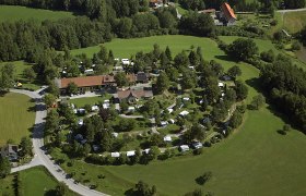 Campinginsel im Grünen, © Reinhard Wallentin Luftaufnahme eines Campingplatzes inmitten grüner Landschaft mit Wohnwagen und Zelten.