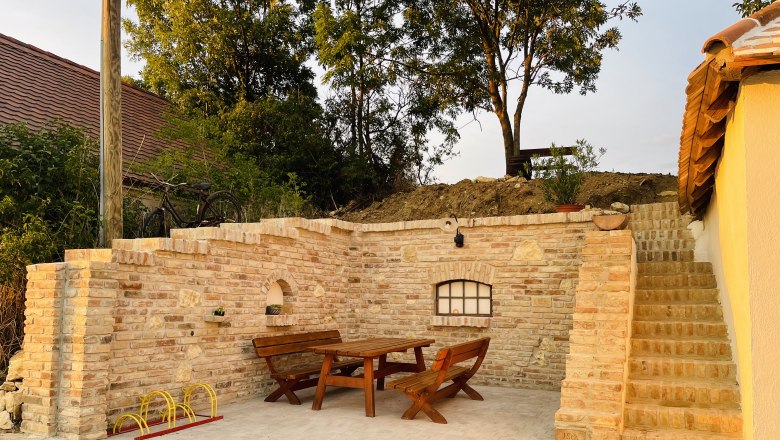 Sitzplatz außen, © Fam. Thürr Gemütlicher Außenbereich mit Holztisch und Bänken, umgeben von Ziegelmauern und Treppe, im Abendlicht.