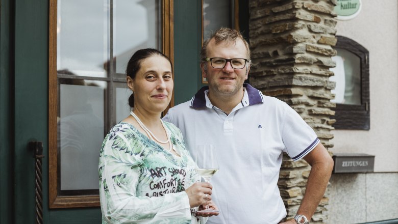 Wirtsleute Martina und Helmut Haas, © Niederösterreich Werbung/David Schreiber Ein Mann und eine Frau stehen lächelnd vor einem Gebäude. Die Frau hält ein Weinglas in der Hand.