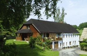 Simandelmühle, © Simandelmühle, Johanna Auer Ein traditionelles Bauernhaus mit Blumen auf dem Balkon, umgeben von grüner Landschaft und Bäumen.