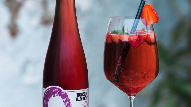 Red Lady vom Bioweingut Jakob, © Bioweingut Jakob Eine Flasche Rotwein mit dem Etikett 'Red Lady' und ein Weinglas mit rotem Getränk und Erdbeere auf einem Holztisch.