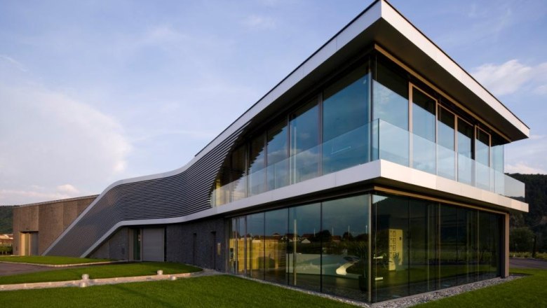 Weingut, © Weinugt FX Pichler Modernes Gebäude mit Glasfassade und geschwungenem Dach, umgeben von grüner Wiese.