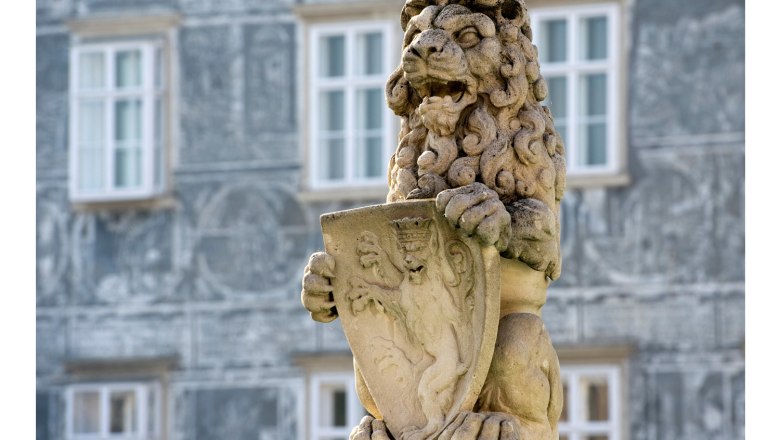 Löwe auf Stadtbrunnen, © Himmel Steinlöwe mit Wappen auf einem Brunnen vor einem Gebäude.