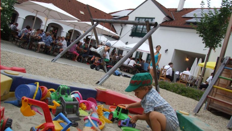 Kinderspielecke, © WalterSchiffmann Ein Kind spielt in einem Sandkasten mit buntem Spielzeug. Im Hintergrund sitzen Menschen an Tischen unter Sonnenschirmen.