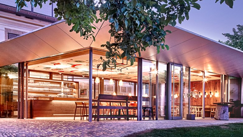 Außenansicht, © Familie Mang Moderne Glasfassade eines Restaurants mit beleuchtetem Innenraum und Bar.