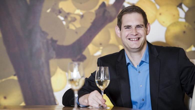 Martin Blaha, © Alfred Kusdat Ein Mann in einem Anzug sitzt an einem Tisch mit zwei Weingläsern vor einem Hintergrund mit Weintrauben.