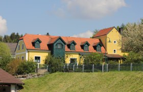 Ferienwohnung Zum alten Forsthaus, © Angela Schmid Gelbes Haus mit rotem Dach und grünen Fensterläden, umgeben von Bäumen und einem Zaun.