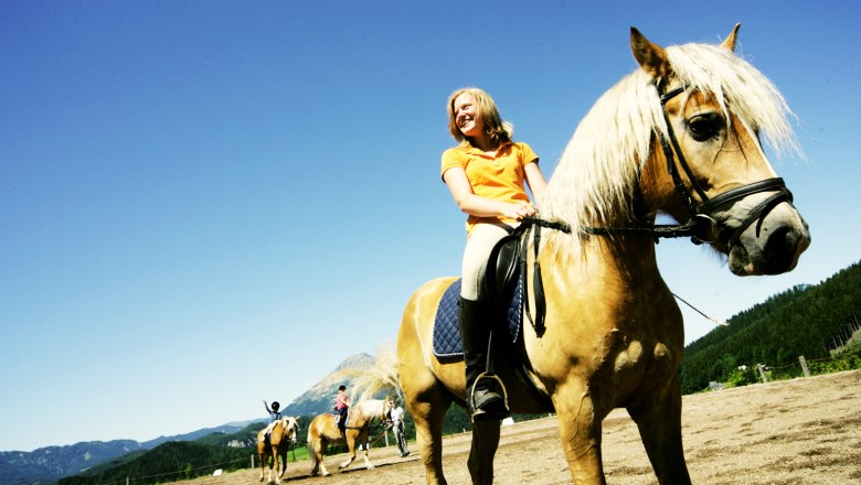 Reiterzentrum Joachimsberg, © Mostviertel Tourismus/Weinfranz.at Eine Frau reitet auf einem braunen Pferd mit blonder Mähne auf einem Reitplatz, umgeben von Bergen und blauem Himmel.