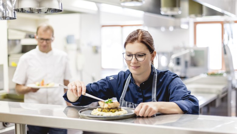 Bringt frischen Wind in die Waldviertler Küche: Tanja Schicker-Mathe, © Niederösterreich Werbung/David Schreiber Köchin in einer Restaurantküche richtet ein Gericht an, während ein Kollege im Hintergrund ein weiteres Gericht hält.