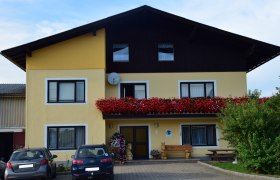 Außenansicht, © Fam. Himmelmayer Außenansicht eines gelben Hauses mit Balkon voller roter Blumen und geparkten Autos davor.