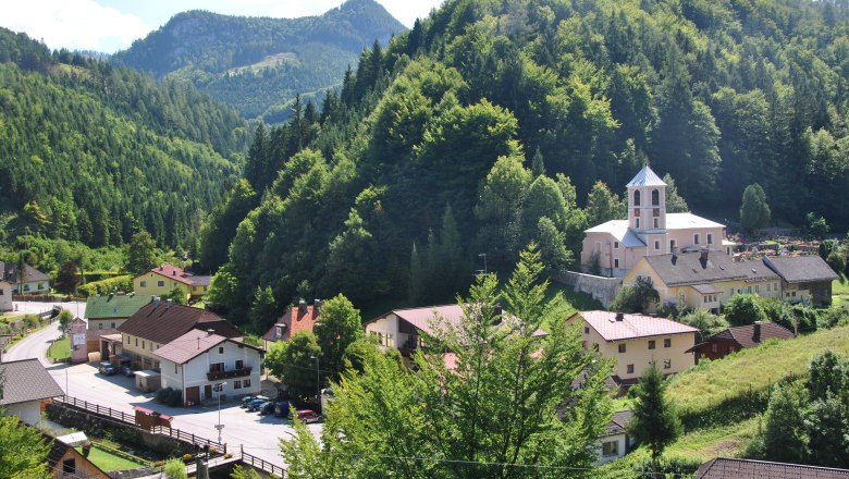 Ortsfoto, © Gemeinde Schwarzenbach/Piel. Kleines Dorf in einer grünen Berglandschaft mit Kirche und Häusern.