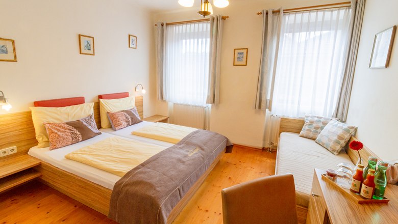 Gästezimmer, © Annemarie Heller Ein gemütliches Gästezimmer mit Doppelbett, Tagesbett, Holzmöbeln und dekorativen Kissen. Helle Vorhänge und Bilder an den Wänden.