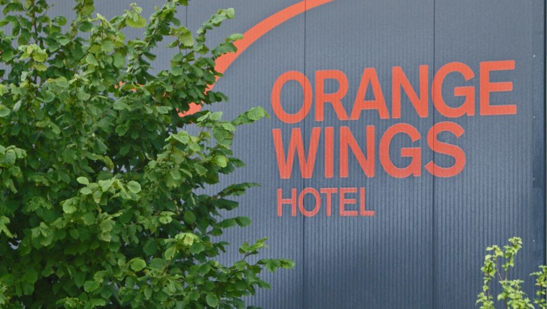 Orange Wings Logo, © Wiener Alpen/Katrin Zeleny Logo des Orange Wings Hotels auf einer Wand mit Pflanzen im Vordergrund.