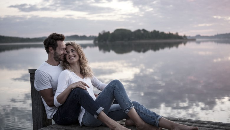 Romantikwochenende im Faulenzerhotel, © Karl Schweighofer Ein Paar sitzt lächelnd auf einem Steg am See bei Sonnenuntergang.