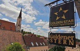 Aussenansicht, © Inhaber Peter Mayer Blick auf ein Schild mit der Aufschrift 'Golden Star Premium Apartments' und 'Goldener Stern' vor einem Kirchturm und Dächern.