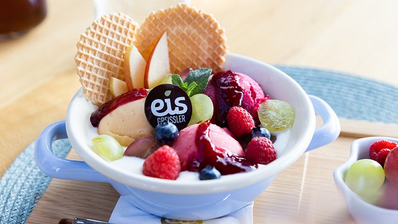 Eis-Greissler Kaffeehaus Joghurtweidling, © Blochberger Eisproduktion GmbH Eisbecher mit Joghurt, Fruchteis, Früchten und Waffeln auf einem Holzbrett.