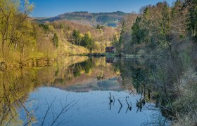 Naturbad Kematen, © zVg Gemeinde Kematen Ein ruhiger See mit Wald und Hügeln im Hintergrund, reflektiert im Wasser.