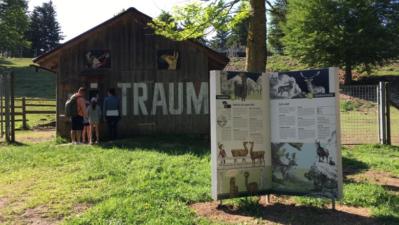 wissensbuecher-6_web, © Naturpark Hohe Wand Eine Familie steht vor einer Holzhütte mit der Aufschrift 'TRAUM'. Daneben eine Infotafel über Tiere in einem grünen Park.
