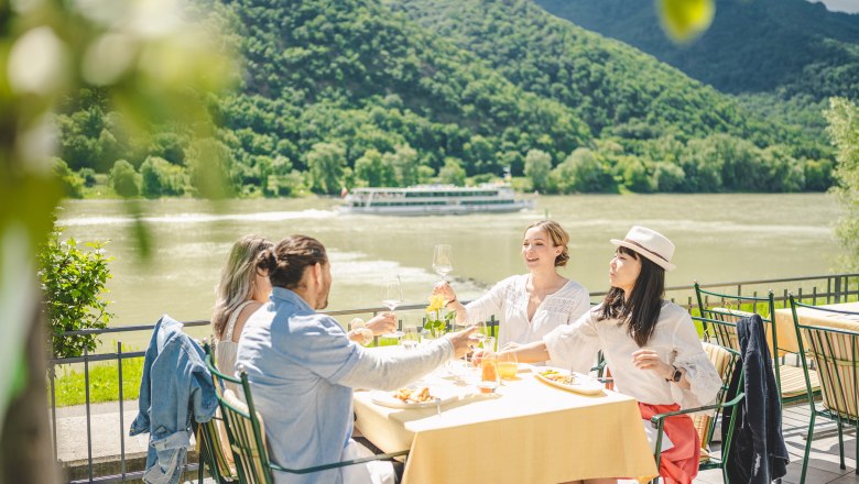 Terrasse mit Ausblick direkt an der Donau, © Die Abbilderei Vier Menschen sitzen an einem schön gedeckten Tisch auf einer Terrasse im HIntergrund die Donau mit einem Schiff