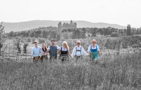 TDR-Hopfeld-Dreikönigshof, © ehnpictures- Johannes Ehn Eine Gruppe von sechs Personen in traditioneller Kleidung steht auf einem Feld mit einer Burg im Hintergrund.