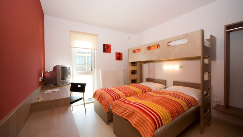 Zimmer, © Nibelungenmotel Pöchlarn Ein modernes Zimmer mit Etagenbett, Schreibtisch, Stuhl und Fernseher. Die Wände sind teils rot gestrichen.