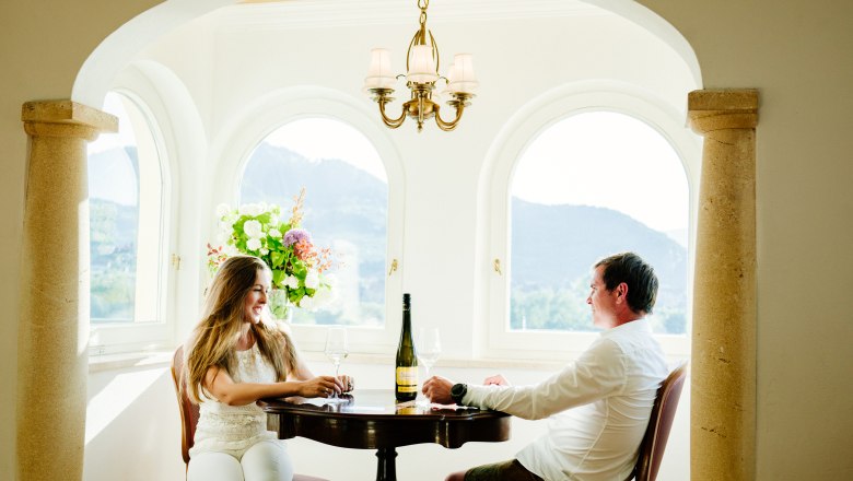 villa_schonthal-29_2, © Julius Hirtzberger Ein Paar sitzt an einem Tisch in einem eleganten Raum mit Rundbogenfenstern und genießt Wein.