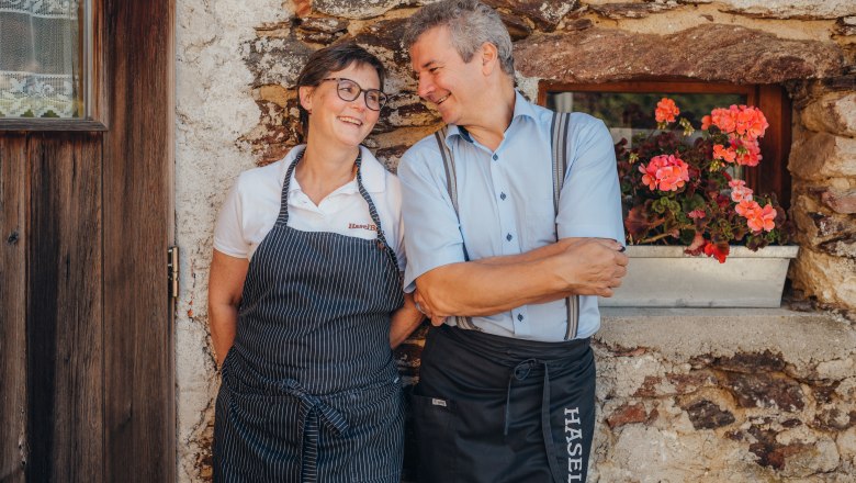 Waltraud & Paul Haselböck, Wirtsleut, © Niederösterreich Werbung/Daniela Führer Ein Mann und eine Frau in Schürzen lehnen lächelnd an einer Steinwand neben einem Fenster mit Blumen.