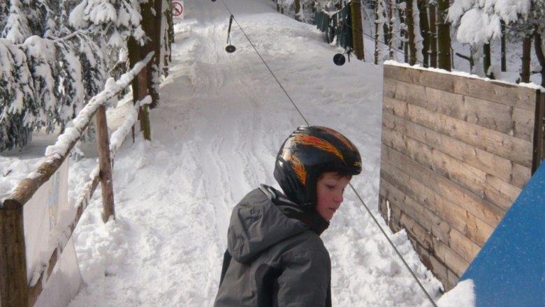 Skigebiet Oberkirchbach, © Bonka GmbH Ein Kind mit Helm steht auf Skiern an einem Schlepplift in einem verschneiten Wald.