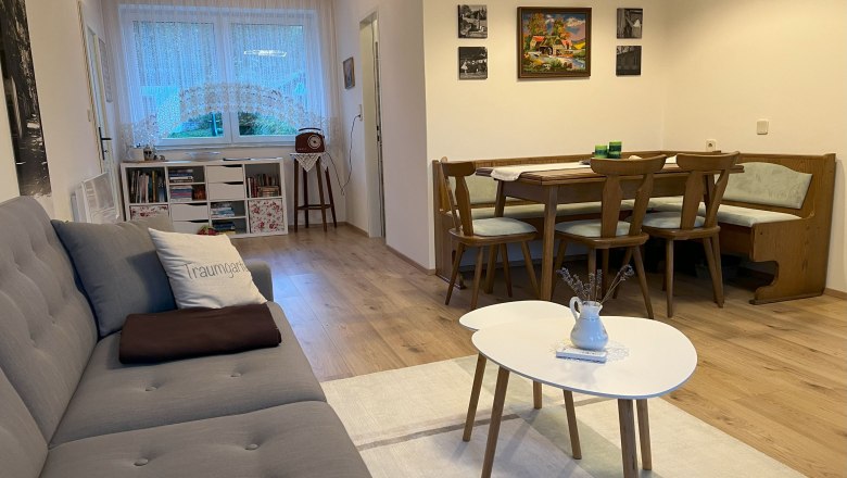 Wohnzimmer, © Waltraud Forstner Gemütliches Wohnzimmer mit grauem Sofa, Esstisch und Stühlen, Bücherregal und Fenster mit Gardinen.