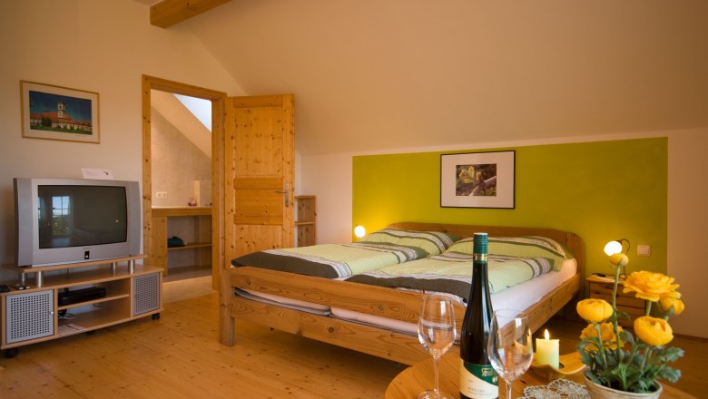 Veltliner - Zimmerfoto, © stradinger Gemütliches Zimmer mit Doppelbett, TV, Weinflasche und Blumen auf Tisch.