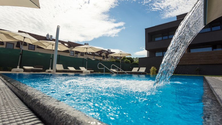 Aussenpool mit Wasserfall, © KASTNER NEW MEDIA - kastner.tv Ein moderner Pool mit Wasserfall, umgeben von Liegestühlen und Sonnenschirmen, bei sonnigem Wetter.