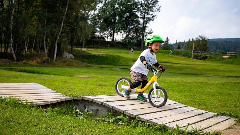 Mini Bikepark Wexl Trails, © Wexl Arena St. Corona am Wechsel Ein Kind mit grünem Helm fährt auf einem Laufrad über eine Holzrampe im Mini Bikepark Wexl Trails.