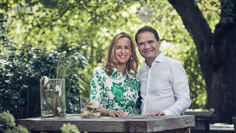 Katharina und Georg Stocker, © Niederösterreich Werbung/Andreas Hofer Ein Mann und eine Frau stehen lächelnd an einem Holztisch im Freien, umgeben von grüner Natur.