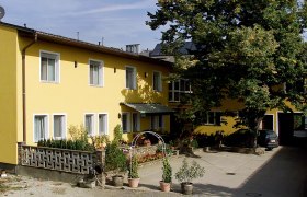 Privatzimmer Familie Holzer, © Fam. Holzer Gelbes Gebäude mit mehreren Fenstern, Pflanzen im Vordergrund und einem großen Baum daneben.