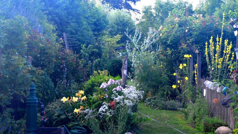 5cf7b1af-5123-422e-be3e-e81130fb86bb_farbe_ausschn, © Familie Mahr Ein üppiger Garten mit verschiedenen Blumen und Büschen, einem grünen Wasserhahn und einer Gießkanne im Vordergrund.