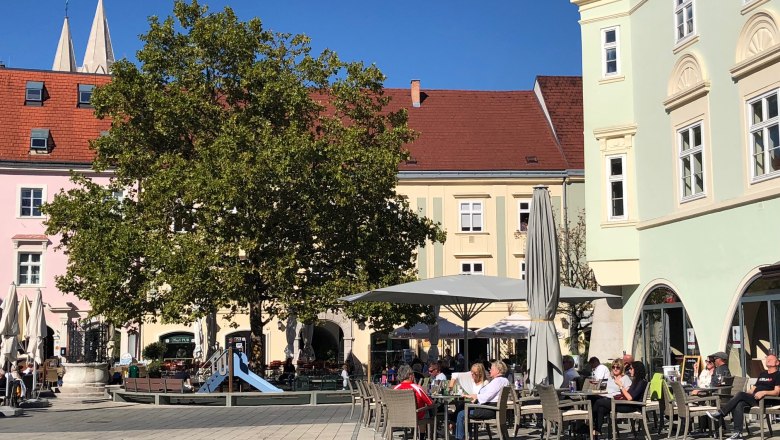 Eterno beim Spielplatz am Hauptplatz, © Birgit Rada-Tomasin Sonniger Hauptplatz mit einem Baum und Spielplatz beim Eterno. Menschen sitzen draußen und genießen das Wetter.