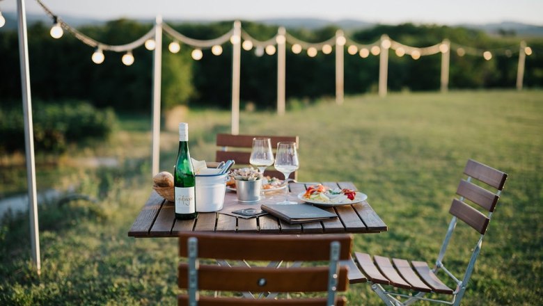 Garten mit Atmosphäre und Aussicht., © Weingut & Weinblick Grafinger Ein Tisch im Freien mit Wein und Snacks, umgeben von Lichterketten und grüner Landschaft.