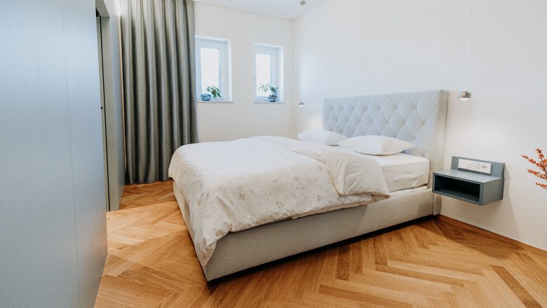 Neu adaptiere Zimmer laden ein, © Der Löffler - Nicole Seiser Modernes Schlafzimmer mit Doppelbett, Parkettboden und grauen Vorhängen.