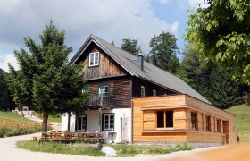 Schutzhaus Vorderötscher, © weinfranz.at Ein traditionelles Holzhaus mit Anbau in einer ländlichen Umgebung, umgeben von Bäumen und Wiesen.
