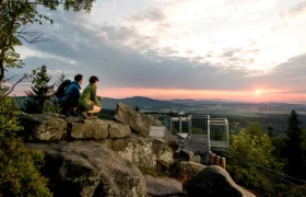 Aussichtsplattform beim Mandelstein, © Studio Kerschbaum Zwei Personen auf einer Felsformation bei Sonnenuntergang mit Blick auf eine Aussichtsplattform und weite Landschaft.