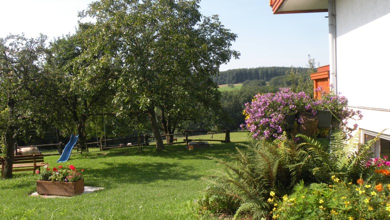 Garten, © Verena Ein grüner Garten mit Bäumen, Blumen und einer blauen Rutsche.