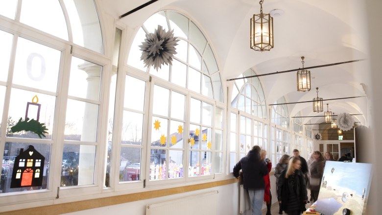 Advent im Schloss Katzelsdorf, © Wiener Alpen/Stefanie Reisenbauer Ein heller Korridor im Schloss mit großen Fenstern und einem Stand am Christkindlmarkt mit Kunst.