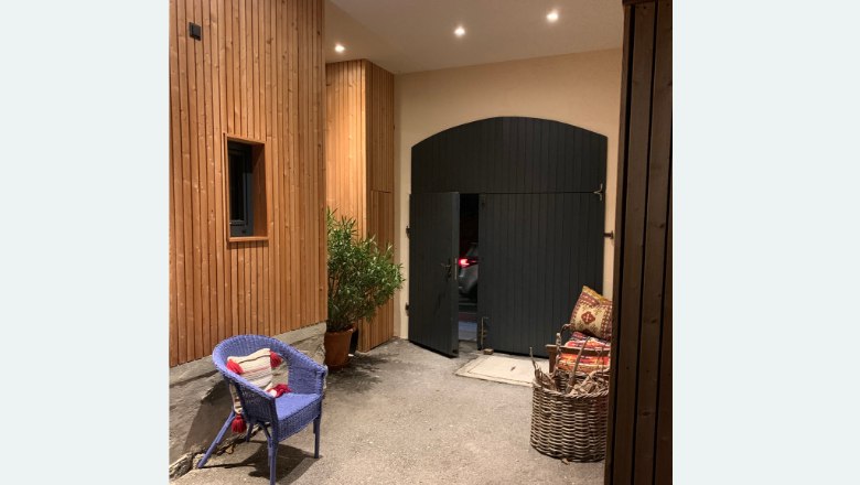 Innenhof Haus Zwei, © Familie Wochesländer Ein Innenhof mit Holzwänden, einem blauen Stuhl, einer Pflanze und einem großen schwarzen Tor.