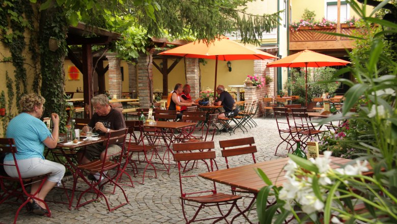 Gausterer, © Gausterer Markus Ein gemütlicher Biergarten mit Holztischen, roten Stühlen und orangefarbenen Sonnenschirmen. Menschen sitzen und essen im Freien.