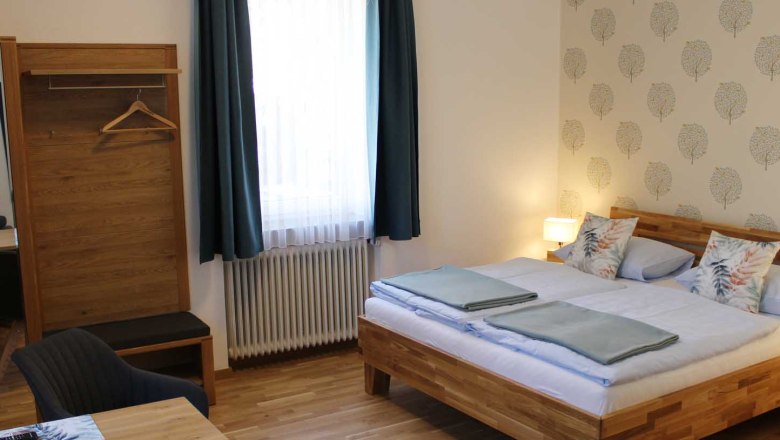 Doppelzimmer, © Magdalena Donhauser Gemütliches Doppelzimmer mit Bett, Kleiderschrank, Tisch, Stuhl und Fenster mit blauen Vorhängen.