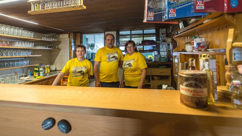 Das Team am Wetterkoglerhaus, © Wiener Alpen, Christian Kremsl Drei Personen in gelben T-Shirts stehen hinter einer Bar in einem rustikalen Raum.