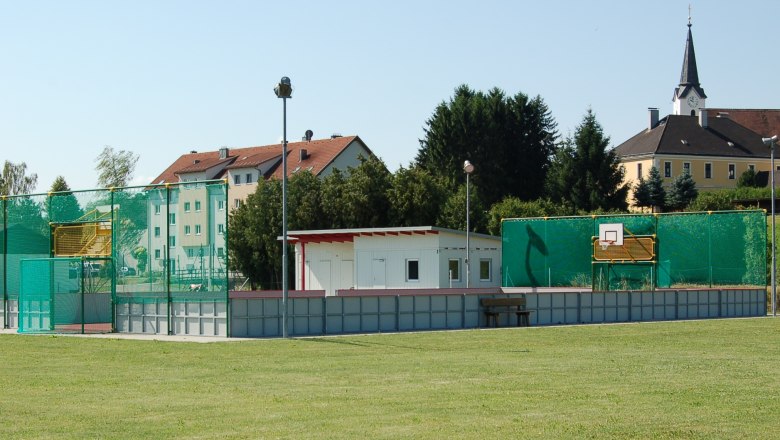 Funcourt und Jugendzentrum, © Gemeinde St. Margarethen Ein Funcourt mit Basketballkorb und grünem Netz, daneben ein kleines Gebäude. Im Hintergrund sind Wohnhäuser und eine Kirche zu sehen.