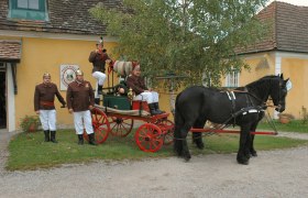 Feuerwehrmuseum Lanzenkirchen, © Gemeinde Lanzenkirchen Historische Feuerwehrkutsche mit Pferd und Feuerwehrleuten in Uniform vor einem gelben Gebäude.