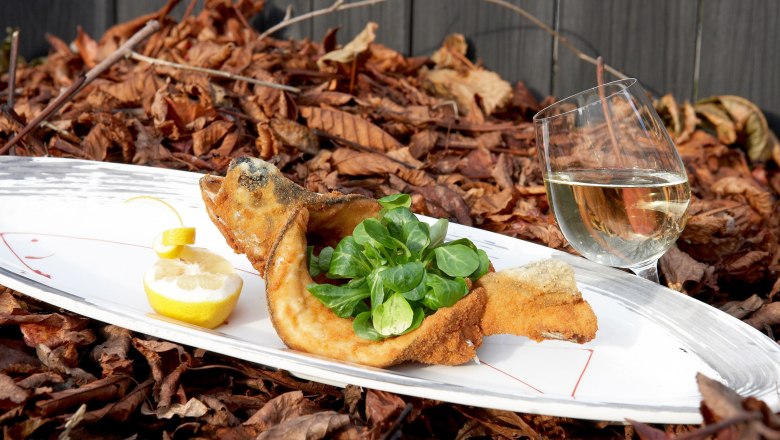 Fisch, © A.Ch. Wulz Gebratener Fisch mit Feldsalat und Zitrone auf einem Teller, daneben ein Glas Weißwein, auf einem Bett aus Herbstlaub.