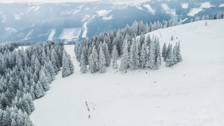 Wechsel Panoramaloipe, © DJI_0557 Verschneite Landschaft mit Langläufern auf einer Loipe, umgeben von schneebedeckten Bäumen und Bergen im Hintergrund.
